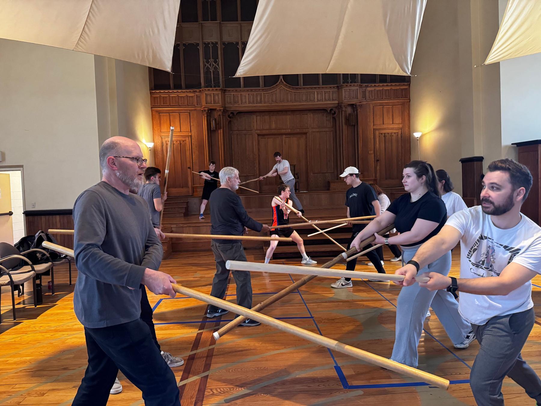 A full WCS Actors Studio class working in pairs with wooden staffs across the rehearsal hall floor